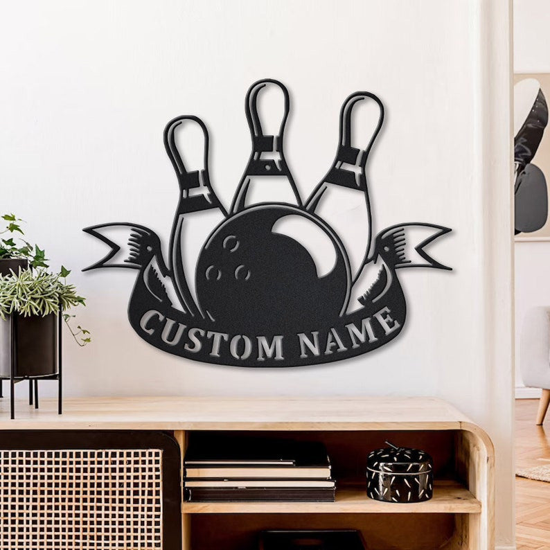 Custom black metal bowling sign mounted on a white wall above a wooden console, featuring a bowling ball, three pins, and a personalized ribbon banner.